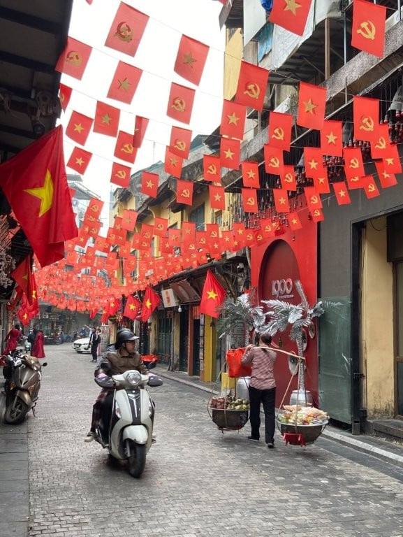 Scooter passing a woman carrying a load of vegetables on flag street