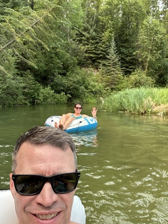 Man taking selfie with a woman floating on the river in the background