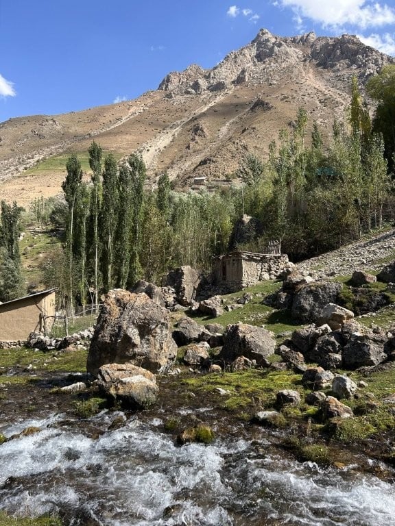 A creek, rocky clearing and adobe house with mountains in the background