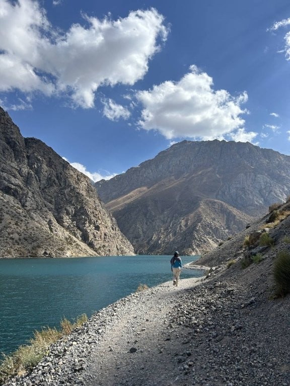 Woman walking along gravel path beside alpine lake in the Fann Mountains